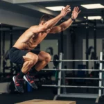 Box Jump Training Guide: Stretch-Shortening Cycle Science, Technique, Landing Mechanics, and Progressive Programming person performing box jump explosive takeoff triple extension arms reaching overhead athletic training gym